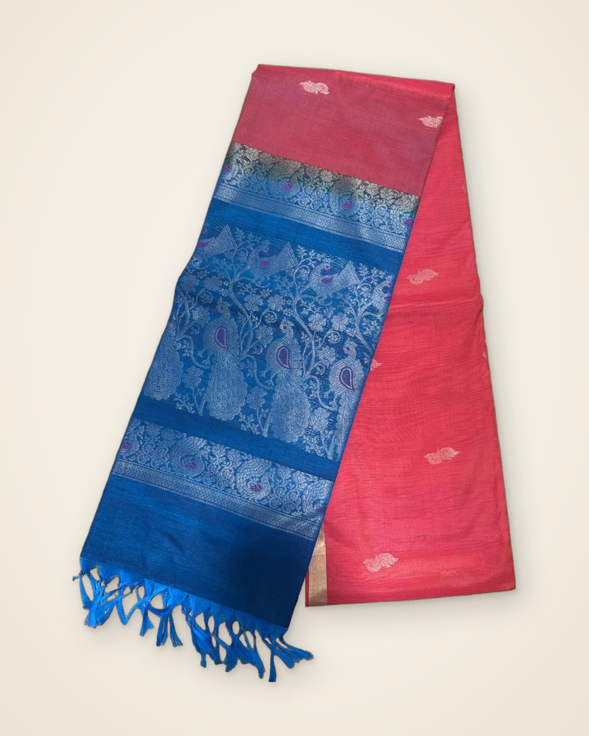 Coral-red saree with a peacock-blue pallu woven with peacocks and vines, folded on a plain ivory backdrop.